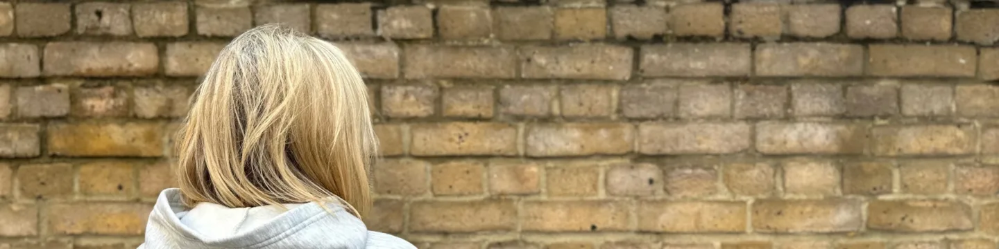 A woman with long blonde hair stands before a brick wall.