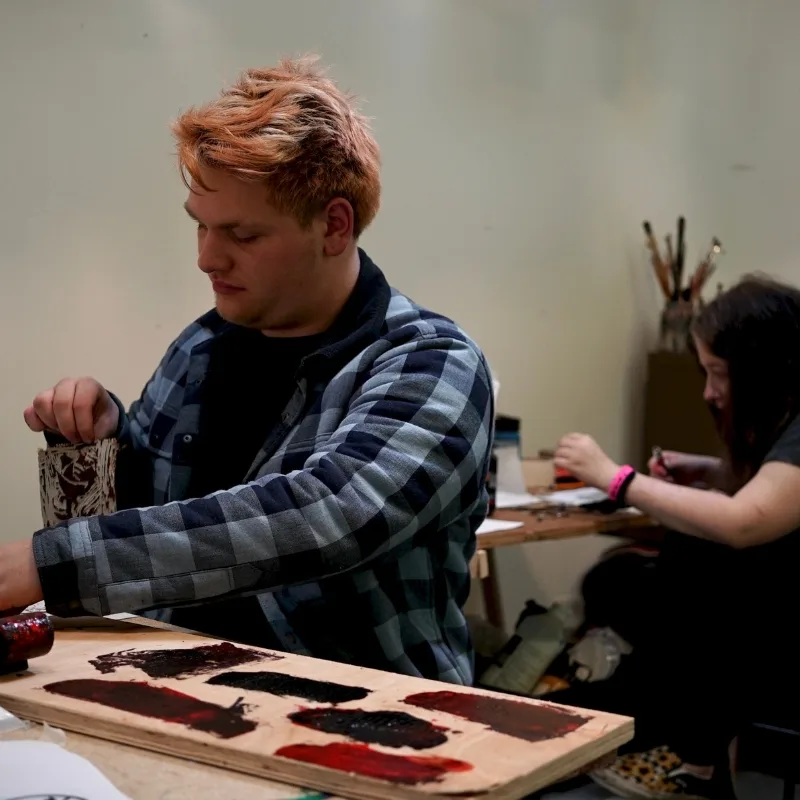 A man and woman are partaking in a screen-printing project, working together in an art studio with tools nearby.
