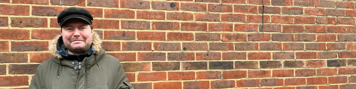 A man in a hat and jacket stands confidently before a brick wall.