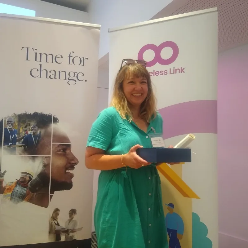 A woman in a green dress smiles while holding a box in front of two banners.