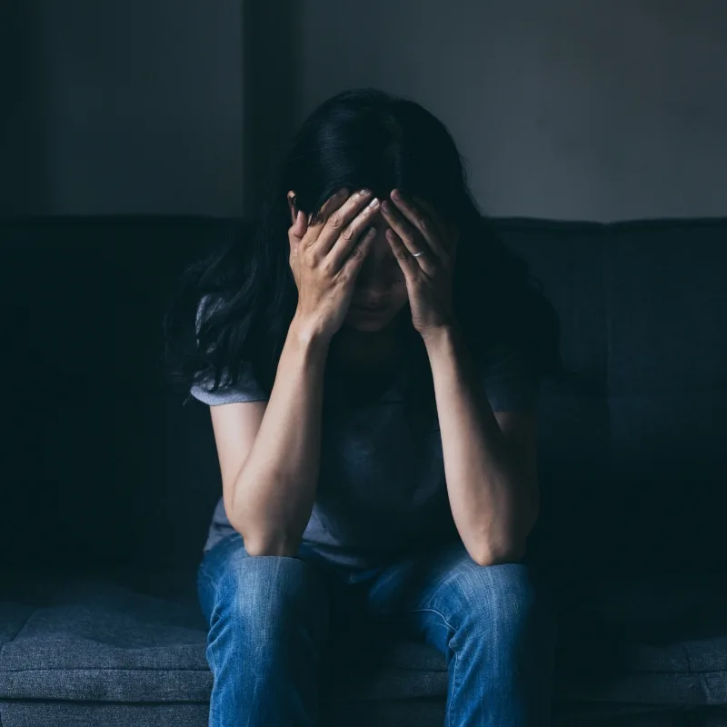 A woman sits on a sofa, covering her face with her hands, conveying deep distress and emotional turmoil.