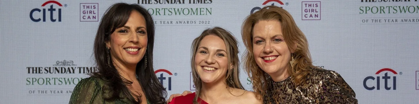Three women proudly stand together, holding an award at an awards ceremony.