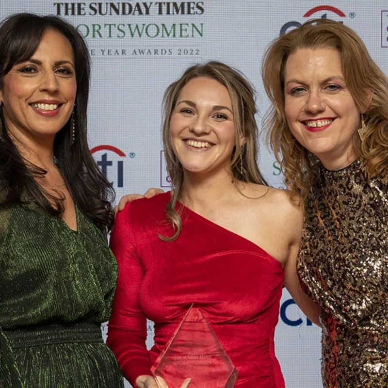 Three women proudly stand together, holding an award at an awards ceremony.