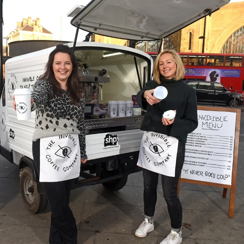 Two women stand in front of a white tuk-tuk holding coffee cups.