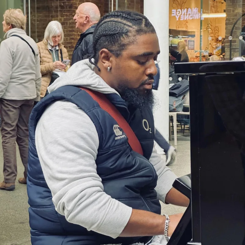 A young man playing a grand piano in a public space, focused on the keys with people passing by in the background.