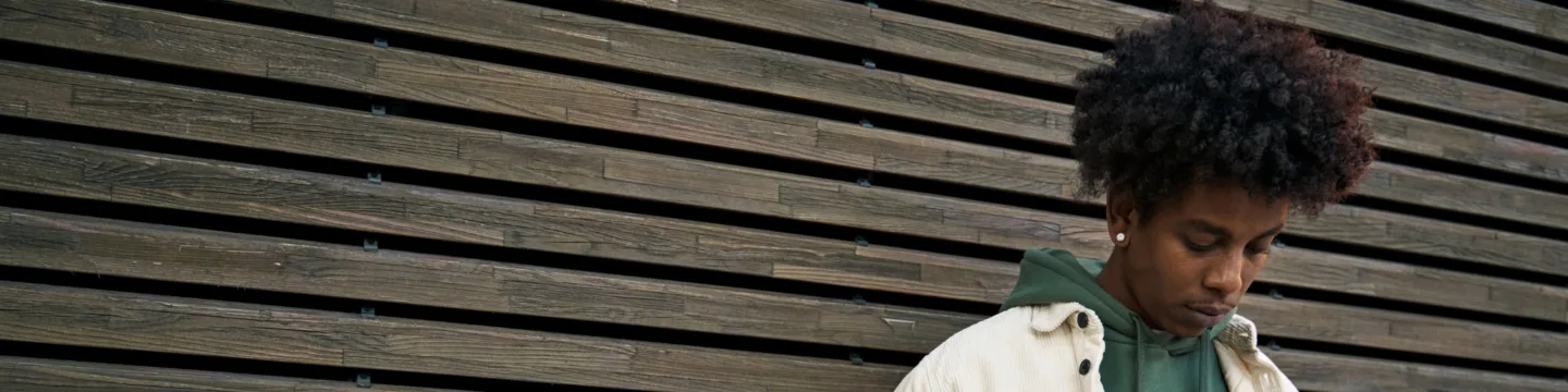 A young man wearing a green hoodie and white jacket poses in front of a wooden wall, looking down at the floor.