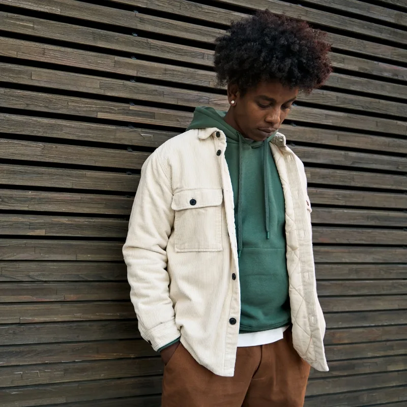 A young man wearing a green hoodie and white jacket poses in front of a wooden wall, looking down at the floor.