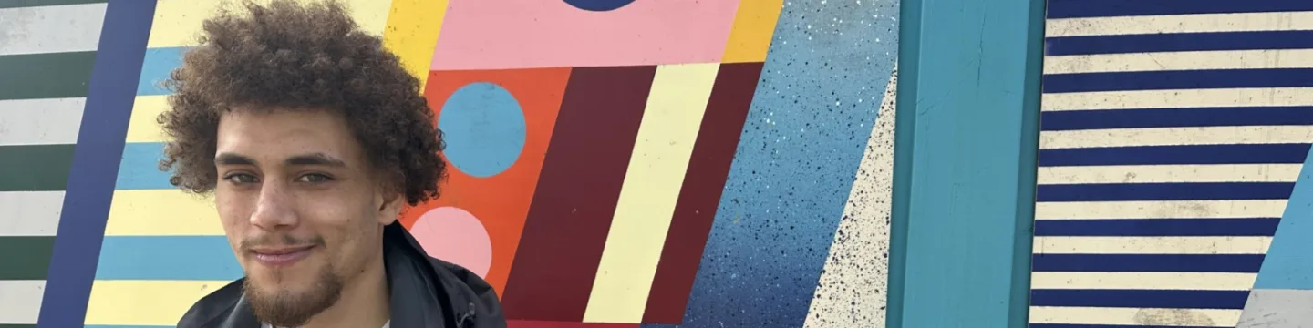 A young man sits relaxed on a bench, smiling in front of a vibrant, colourful wall.