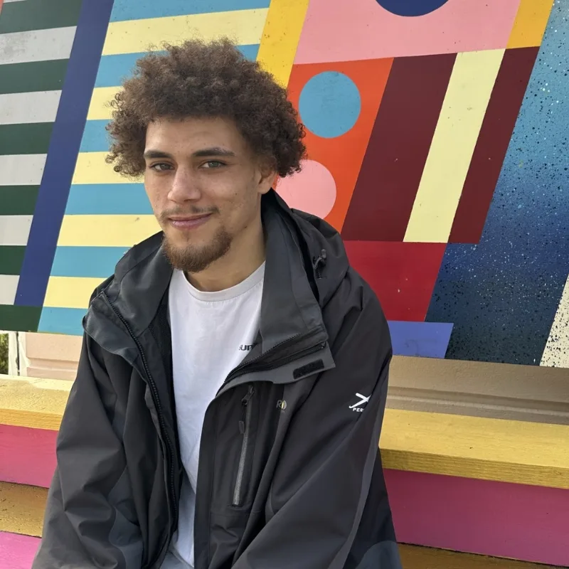 A young man sits relaxed on a bench, smiling in front of a vibrant, colourful wall.