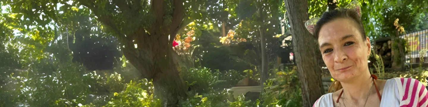 A woman with brown hair smiles in a peaceful park setting, with plants and trees in the background.