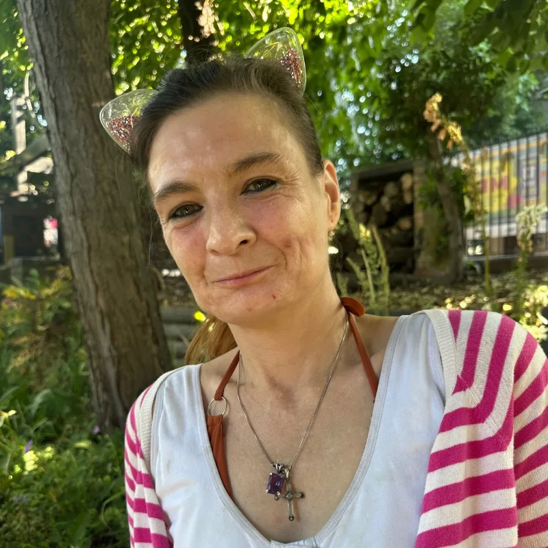 A woman with brown hair smiles in a peaceful park setting, with plants and trees in the background.