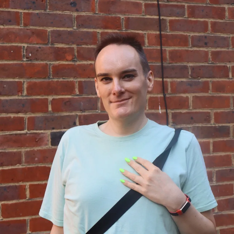 A smiling man with green-painted nails stands confidently in front of a brick wall.