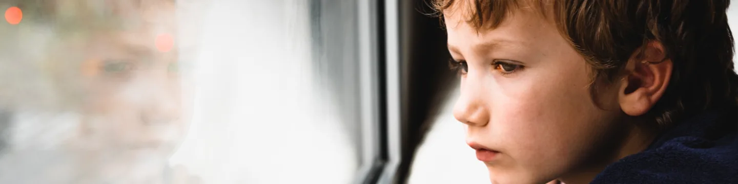 A young boy gazes out of a window with a sad, distant expression.