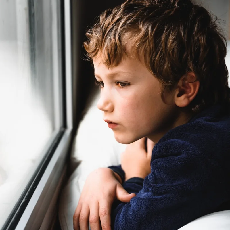 A young boy gazes out of a window with a sad, distant expression.