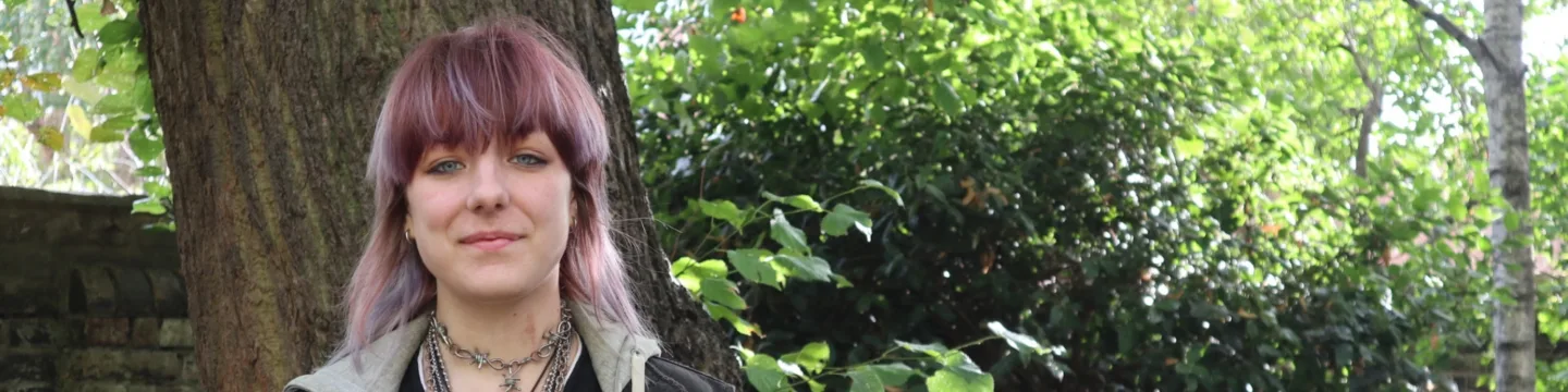 A person with purple hair stands beside a tree in a garden, wearing layered necklaces and at ease.