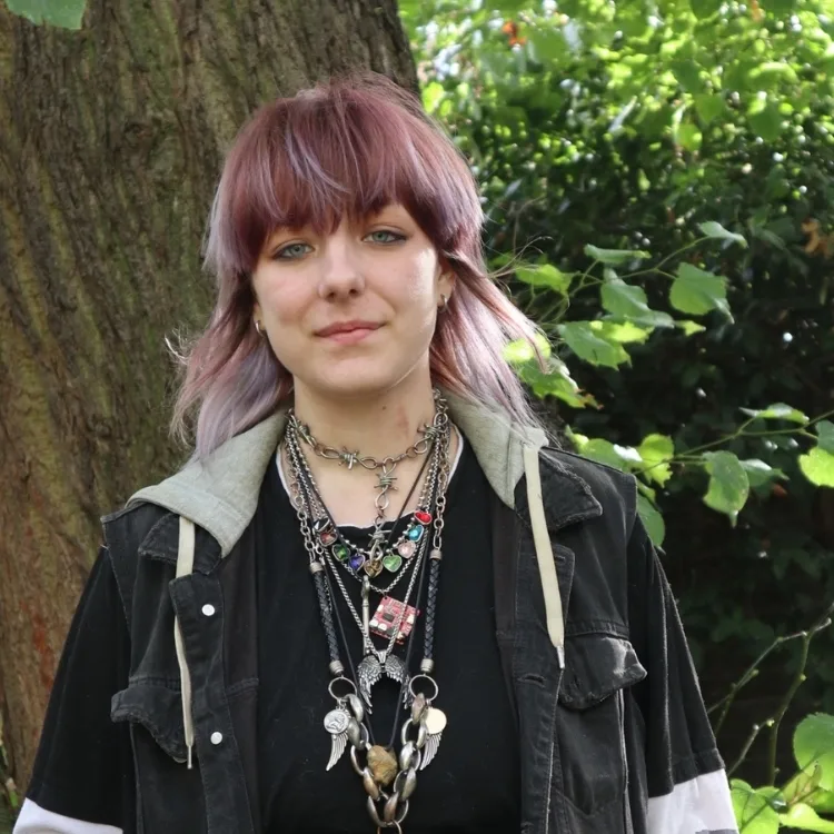 A person with purple hair stands beside a tree in a garden, wearing layered necklaces and at ease.