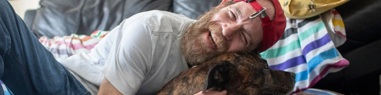 A bearded man in a red cap joyfully hugs a dog on the floor