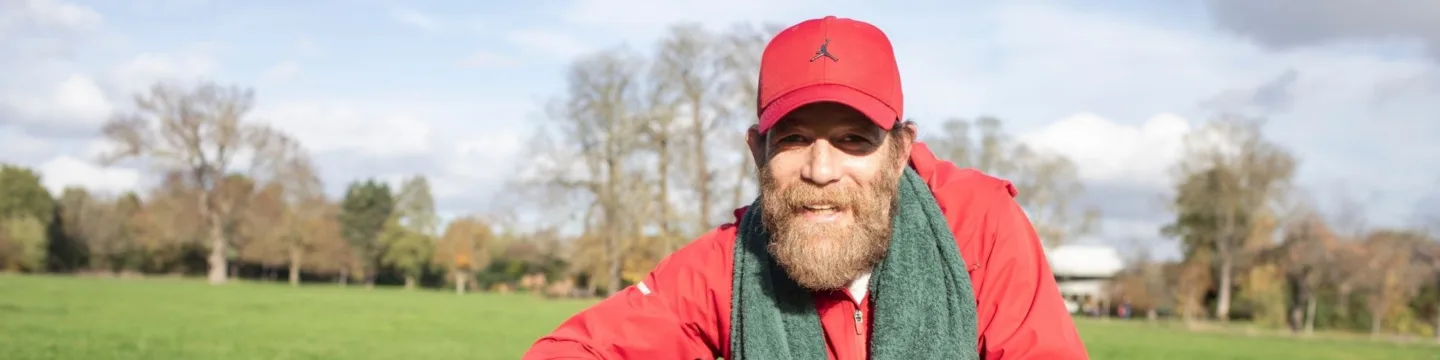 Bearded man in red cap and jacket smiles broadly in a sunny park. Green field and trees in the background