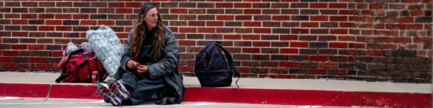 Woman who is rough sleeping sits on pavement with red brick wall behind her