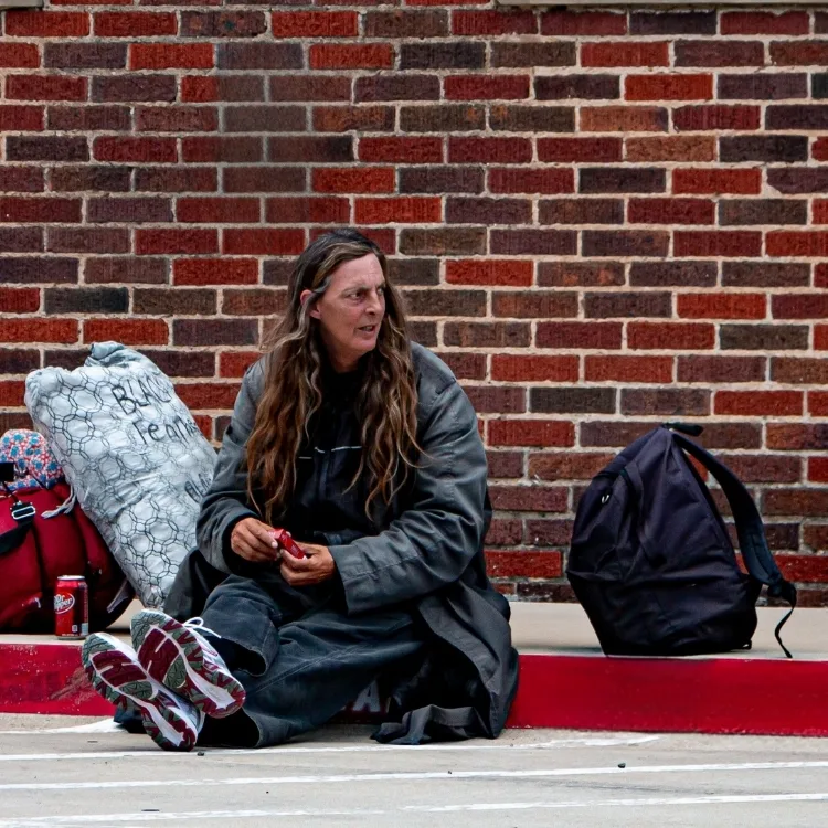 Woman who is rough sleeping sits on pavement with red brick wall behind her