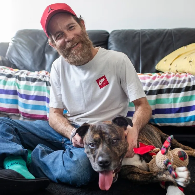 Smiling man in a red cap and white shirt sits on the floor beside a happy brown dog with a toy.