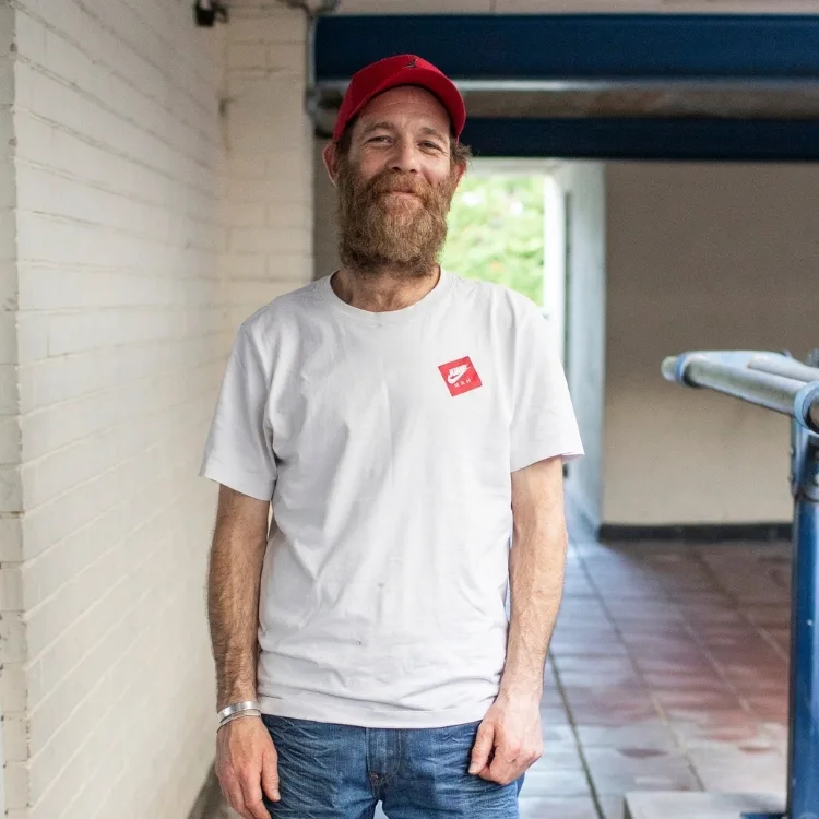A smiling man with a beard wearing a white T-shirt, red cap, and jeans stands in a hallway with white brick walls.