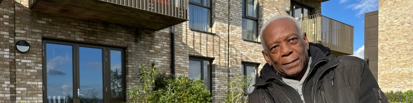 Elderly man with short white hair wearing a black jacket smiles warmly in the sunlight. He's standing against a brick wall with green plants.