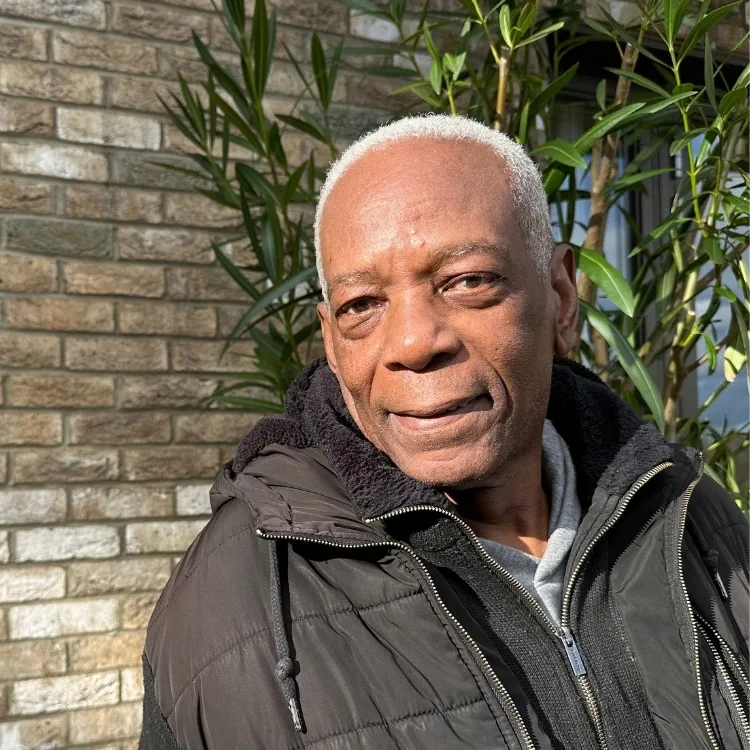 Elderly man with short white hair wearing a black jacket smiles warmly in the sunlight. He's standing against a brick wall with green plants.
