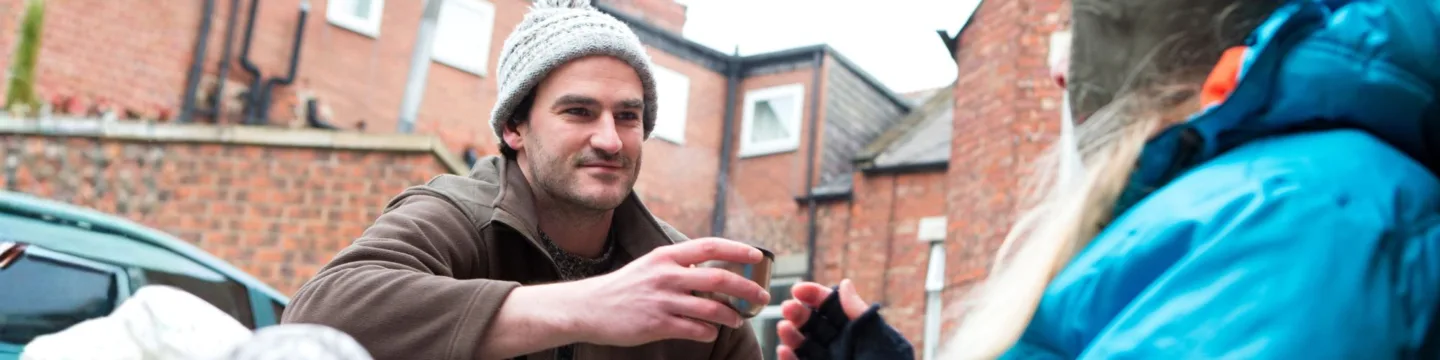 Young man with beanie giving a person rough sleeping a hot drink