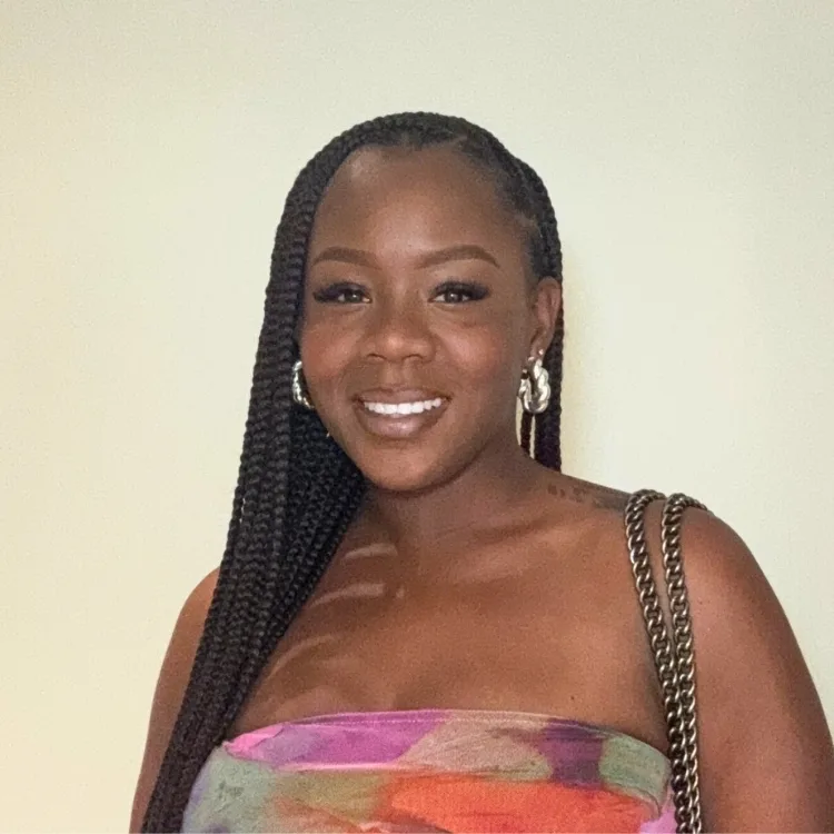 A woman with long braids stands in front of a plain wall, looking relaxed.