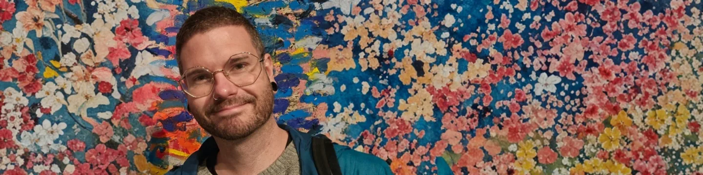 A man with short hair, glasses, and a beard stands in front of a colorful, abstract painted wall, smiling at the camera.