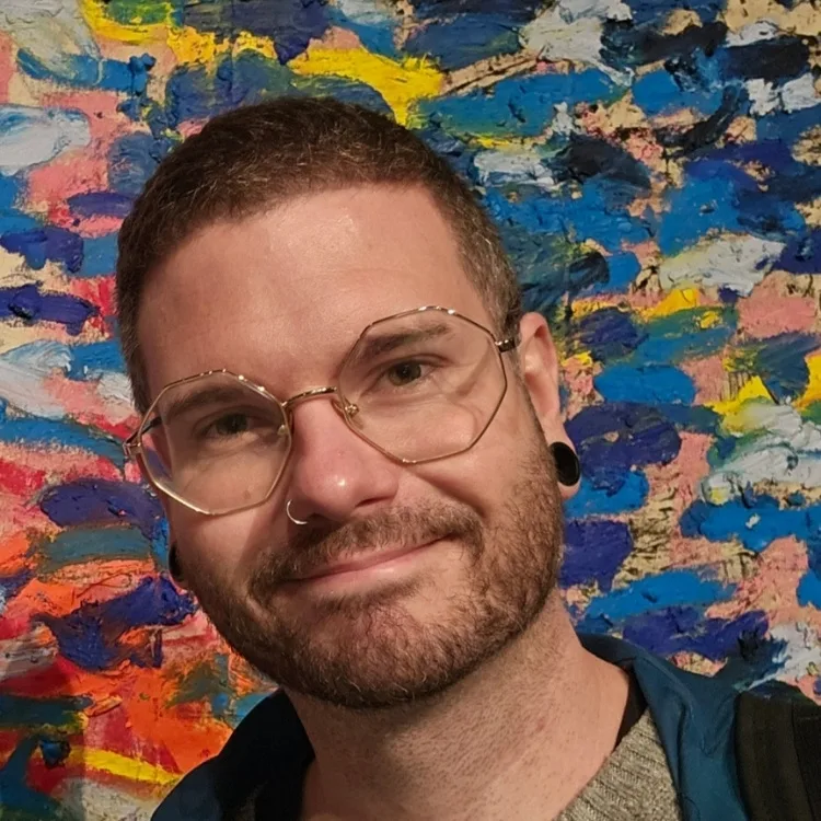 A man with short hair, glasses, and a beard stands in front of a colorful, abstract painted wall, smiling at the camera.
