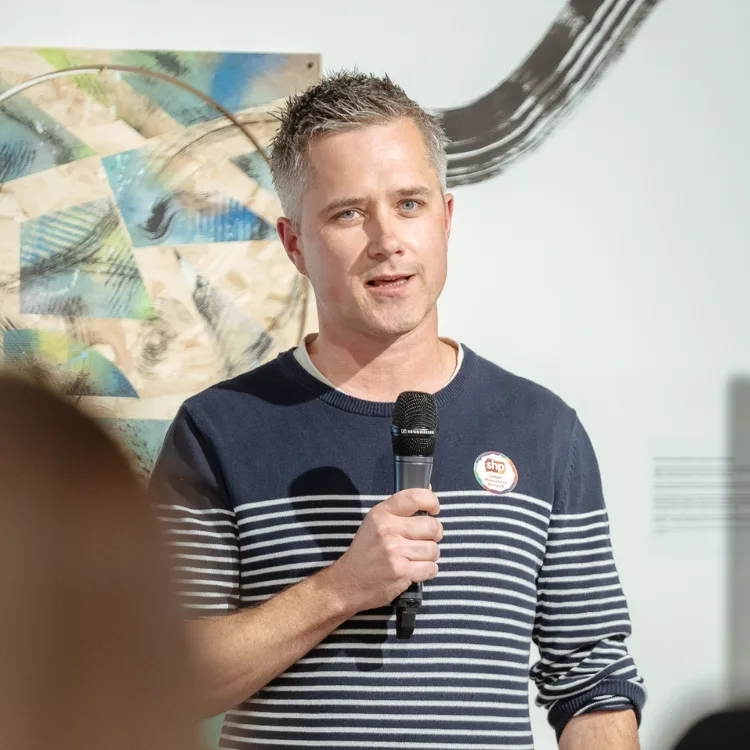 A man with short hair, wearing a striped sweater, speaks into a microphone to an audience.