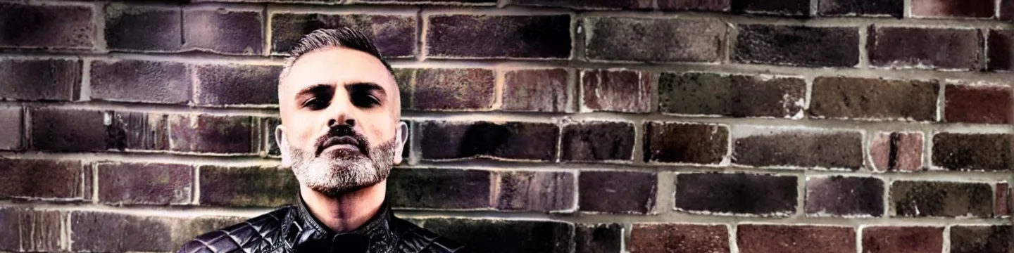 A man with a beard looks at the camera, standing against a brick wall.