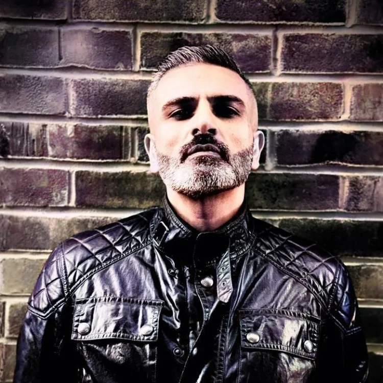 A man with a beard looks at the camera, standing against a brick wall.