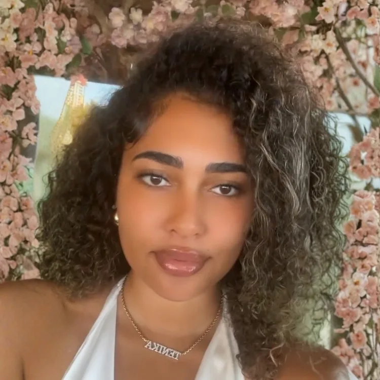 Young woman with curly hair and a white top stands before a backdrop of flowers