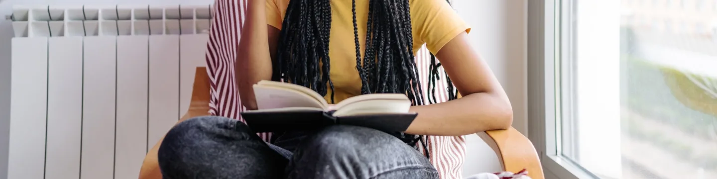 A person with long braids, wearing a yellow top and jeans, sits in a chair while reading a book near a bright window