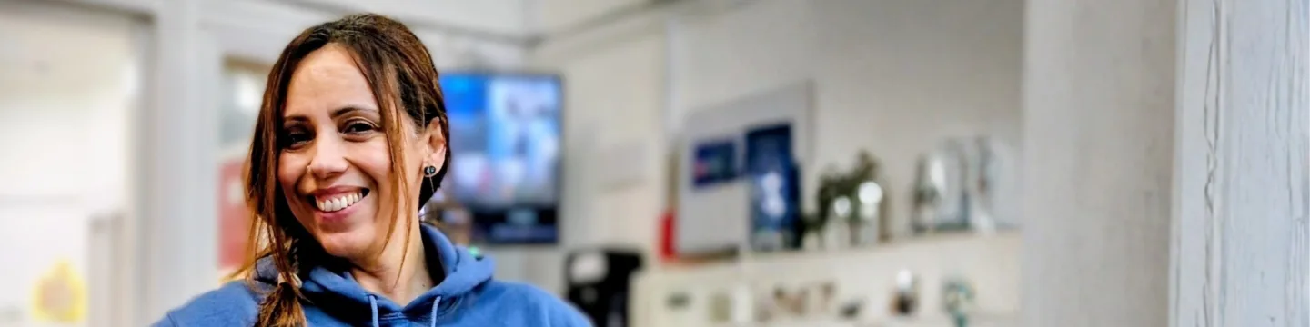 Woman smiling and wearing a blue hoodie, standing indoors with a blurred background.